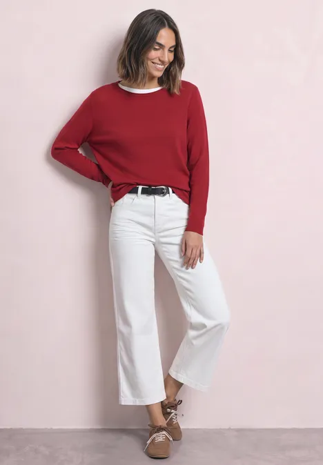 Frau in rotem Pullover, weißen Culotte-Jeans und braunen Sandalen vor rosa Hintergrund.