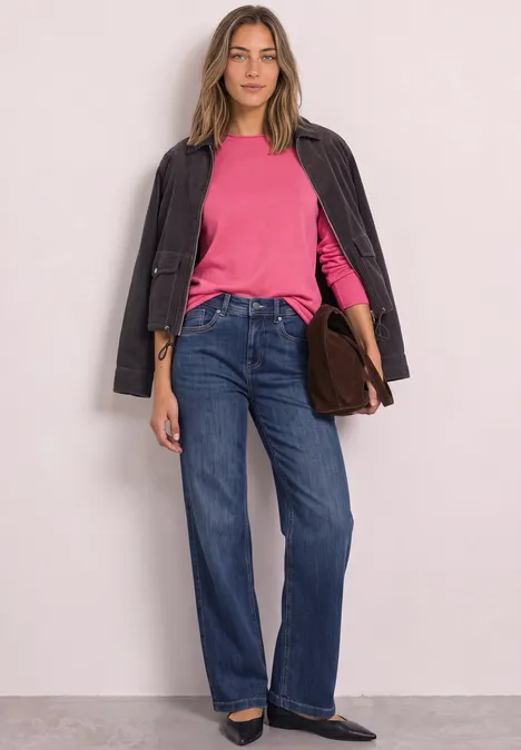 Frau in Stehpose trägt pinkes Oberteil, schwarze Jacke, weite blaue Jeans und schwarze Schuhe, hält eine braune Tasche.