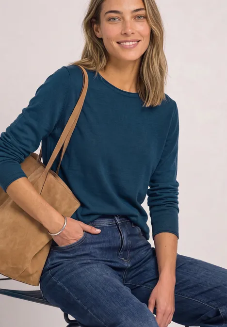 Pullover mit Rundhals und Rollkanten blue petrol Frau in tealfarbenem Langarmshirt und Jeans sitzt lässig mit brauner Wildledertasche und lächelt.
