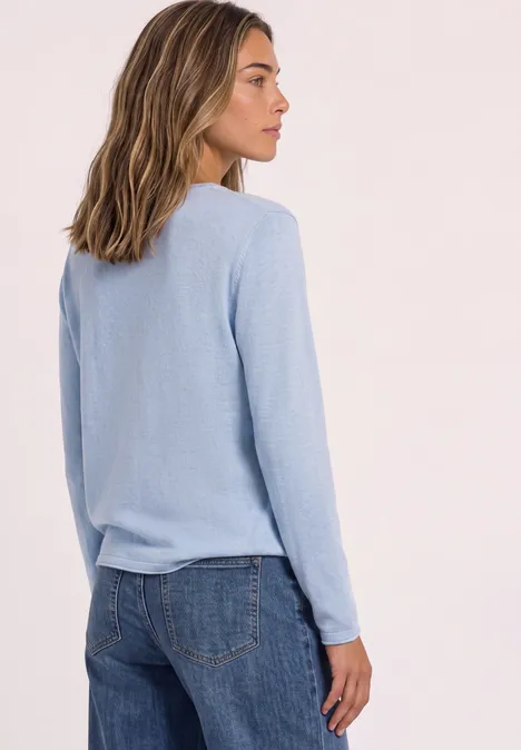 Frau in hellblauem Pullover und Jeans, Seitenansicht vor neutralem Hintergrund.