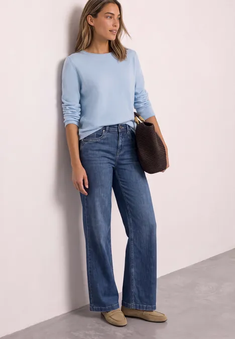 Frau in hellblauem Pullover, dunklen weiten Jeans und Hausschuhen lehnt lässig an heller Wand mit Tasche.