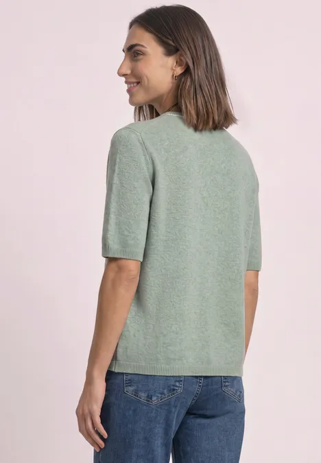 Frau von hinten in hellgrünem Kurzarm-Pullover und blauen Jeans, leicht zur Seite blickend vor hellem Hintergrund.