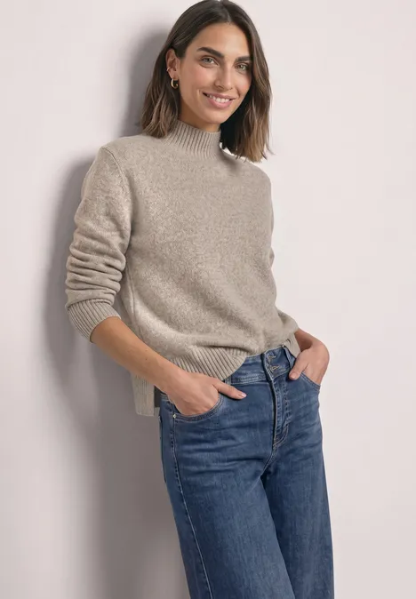 Une femme souriante en pull beige et jean bleu, posant de manière décontractée les mains dans les poches.