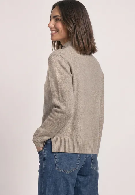Une femme en pull beige et jean bleu vue de profil, souriante et regardant vers la gauche.