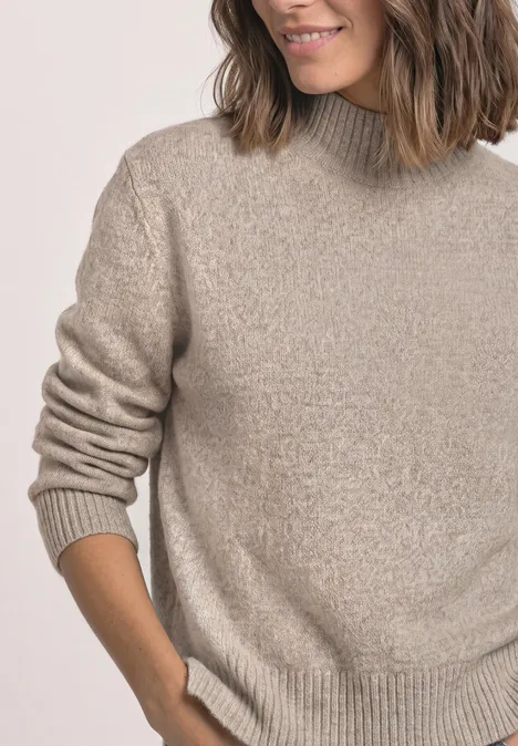 Une femme souriante porte un pull beige à col roulé et manches longues.