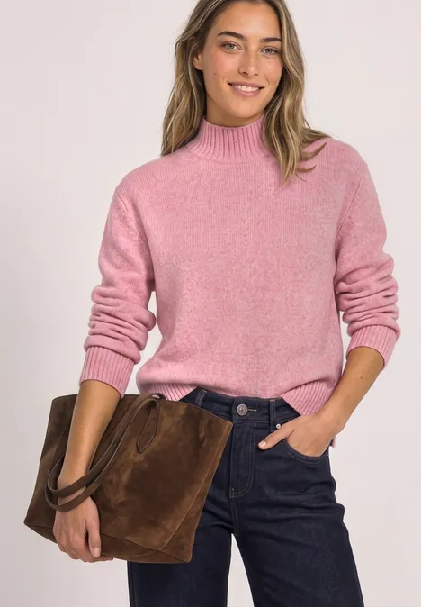 Frau mit rosa Pullover und dunklen Jeans hält eine braune Wildleder-Tasche und lächelt.