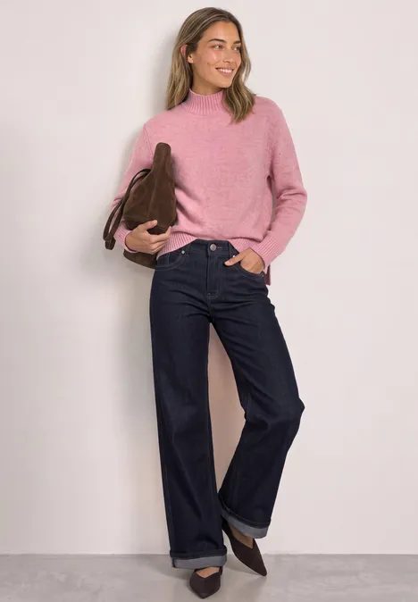 Frau in pinkem Pullover, weiten blauen Jeans und braunen Schuhen hält Handtasche und lächelt vor hellem Hintergrund.