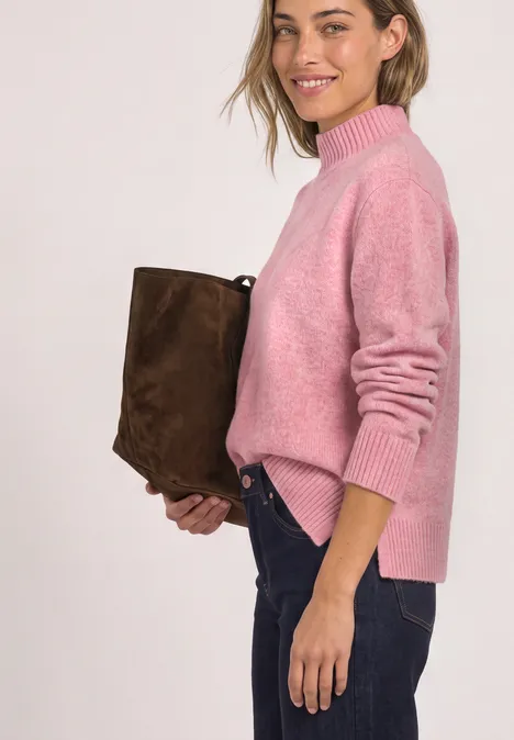 Frau in rosa Pullover und Jeans hält eine braune Tasche und lächelt seitlich.