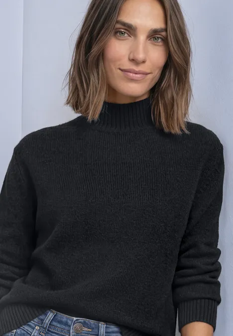 Schwarzer Rollkragenpullover, Jeans, Frau mit schulterlangen braunen Haaren vor hellem Hintergrund.
