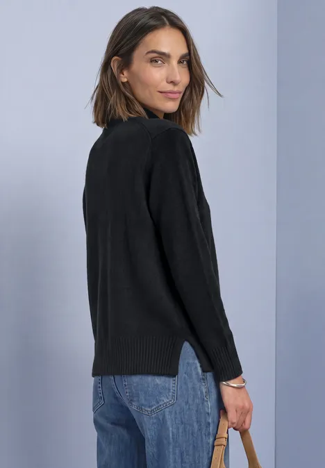 Frau in schwarzem Pullover und Jeans schaut über die Schulter vor hellviolettem Hintergrund.