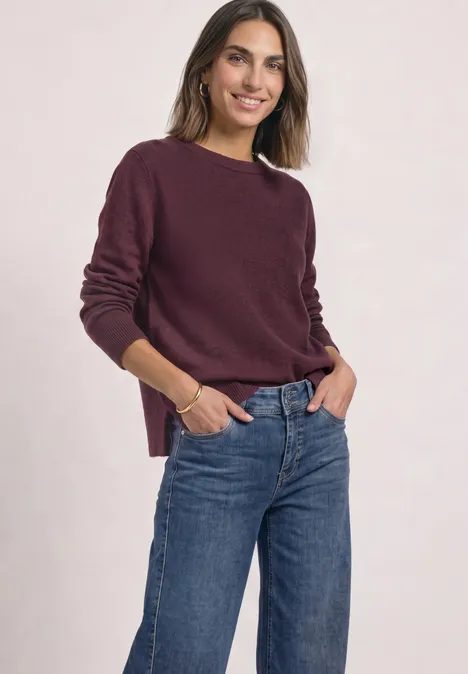 Une femme souriante porte un pull bordeaux et un jean bleu.