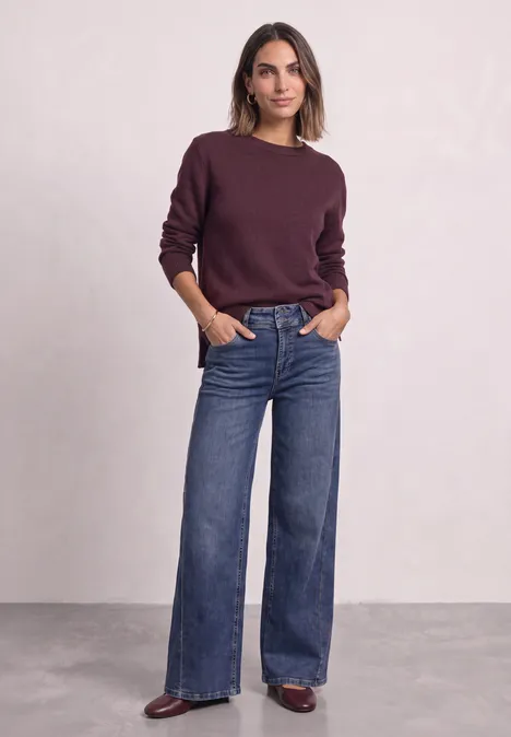 Femme en pull bordeaux et jean large, debout de manière décontractée les mains dans les poches.
