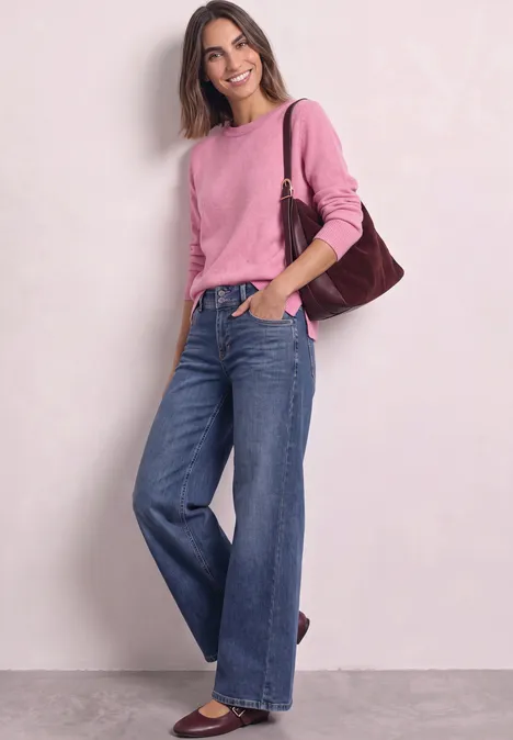 Junge Frau steht lächelnd in weitem Jeanshosen und rosa Pullover, mit brauner Schultertasche.