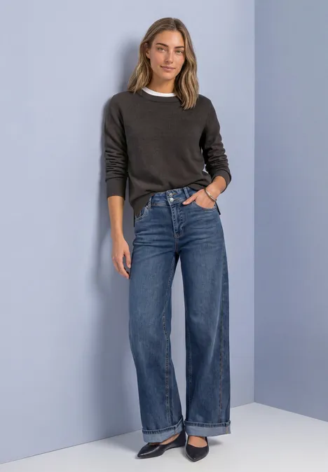 Jeune femme en pull gris, t-shirt blanc et jean large pose décontractée devant un mur bicolore.