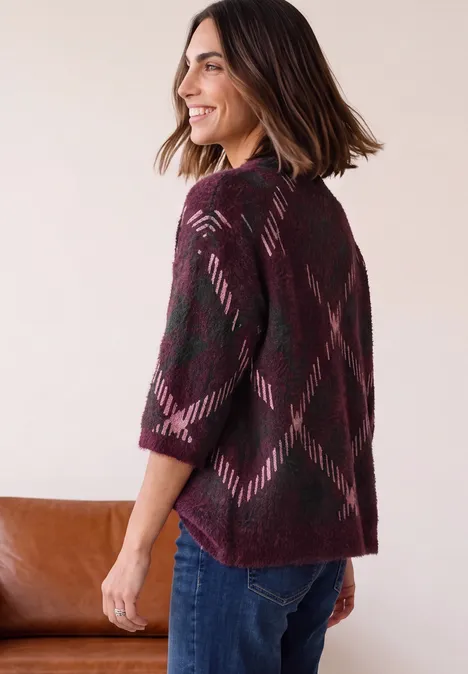 Wzorzysty sweter z rękawem 3/4 i okrągłym dekoltem rustic rouge Kobieta z profilu w bordowej dzianinowej narzutce z geometrycznym wzorem i dżinsach.