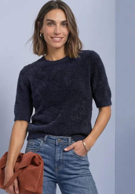 Vrouw lacht in donkerblauwe, pluizige korte trui en jeans, houdt bruin tasje vast voor een lichtblauwe achtergrond.
