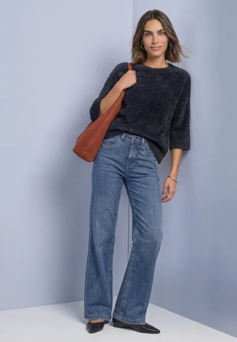 Vrouw met donkergrijze trui, high-waist flared jeans, bruine schoudertas en zwarte schoenen tegen neutrale achtergrond.