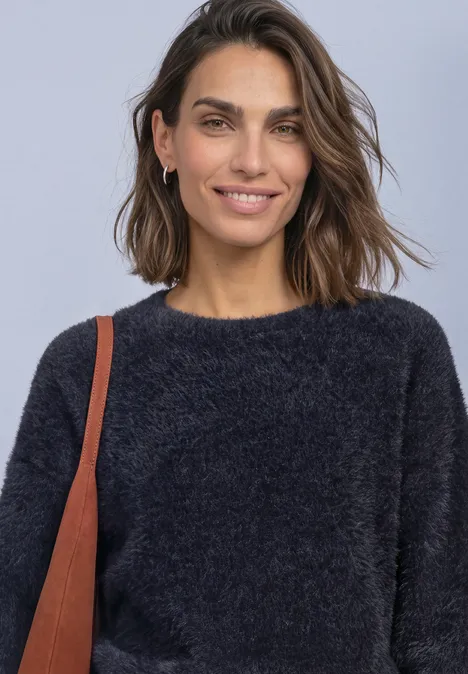 Een vrouw met kort bruin haar, glimlacht, draagt een zwarte, pluizige trui en een schoudertas.