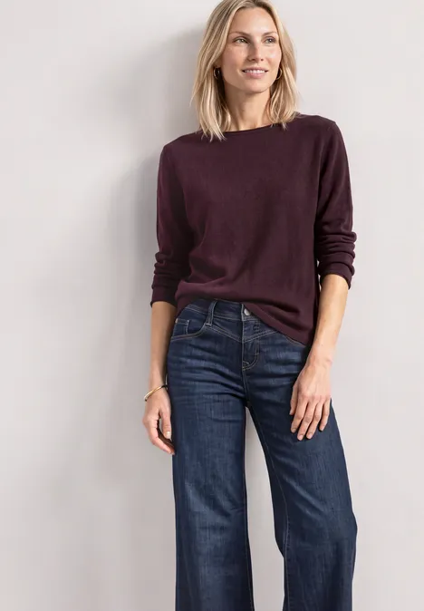 Frau in weinrotem Pullover und weitem Jeans vor neutraler Wand.