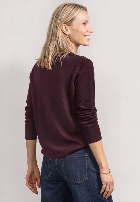 Blonde Frau in dunkelrotem Pullover und Jeans, sieht seitlich weg und lächelt.