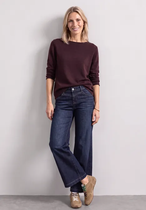 Eine Frau mit blondem Haar trägt Burgunder-Pullover, weite Jeans und Sneaker vor neutralem Hintergrund.