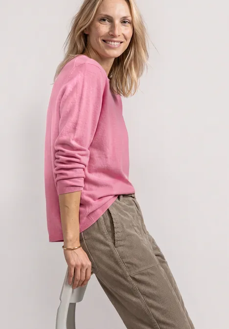 Trui met boothals en omgeslagen randen warm blush mel. Vrouw in roze trui en beige broek, zijwaarts staand en glimlachend voor een lichte achtergrond.