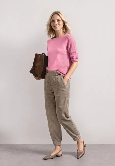 Trui met boothals en omgeslagen randen warm blush mel. Vrouw in roze trui, kaki cargobroek en bruine clutch staat voor een lichte achtergrond.