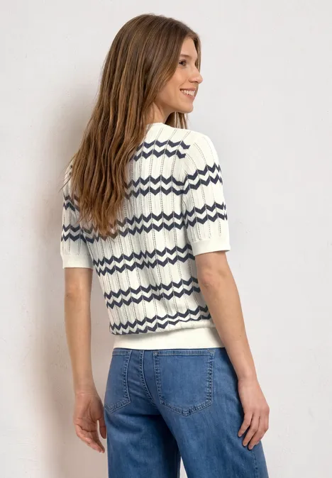 Rückansicht einer Person in blauen Jeans und weißem Shirt mit dunkelblauen Zickzackstreifen vor hellem Hintergrund.