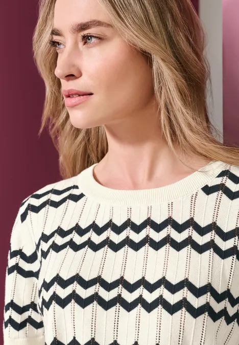 Frau in hellem gestrickten Pullover mit dunklem Zickzackmuster, Blick seitlich nach rechts.