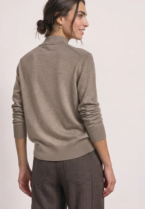 Basic sweter z golfem sanded amber mel. Kobieta od tyłu w beżowym swetrze i brązowych spodniach, ciemne włosy w kok, patrzy w bok.