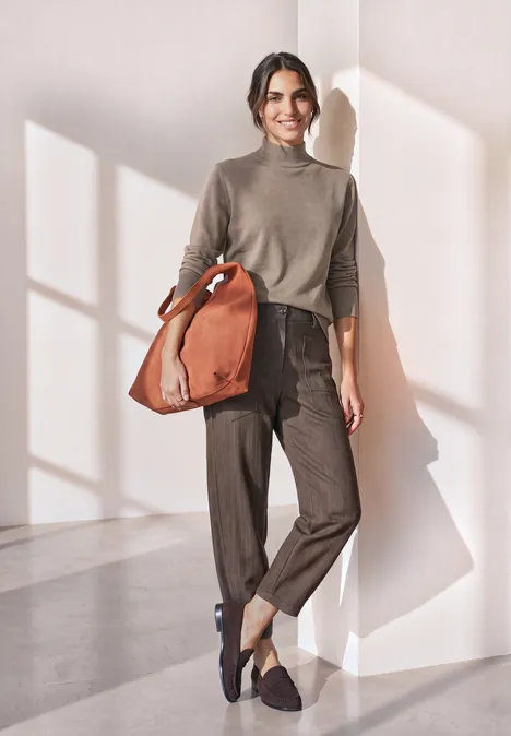 Basic sweter z golfem sanded amber mel. Kobieta w casual stroju z brązową torbą stoi w jasnym wnętrzu.