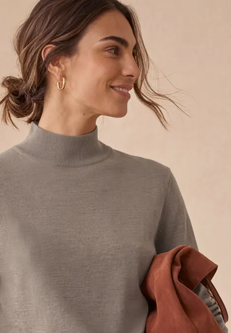 Basic sweter z golfem sanded amber mel. Młoda kobieta w szarym golfie trzyma brązową kurtkę, uśmiecha się z profilu na jasnym tle.