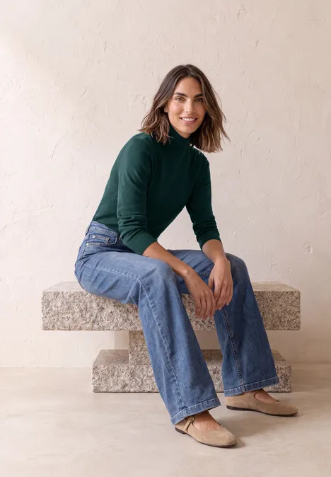 Basic sweter z golfem shadow teal Kobieta siedzi na kamiennej ławce, ma na sobie zielony golf, dżinsy i baleriny.