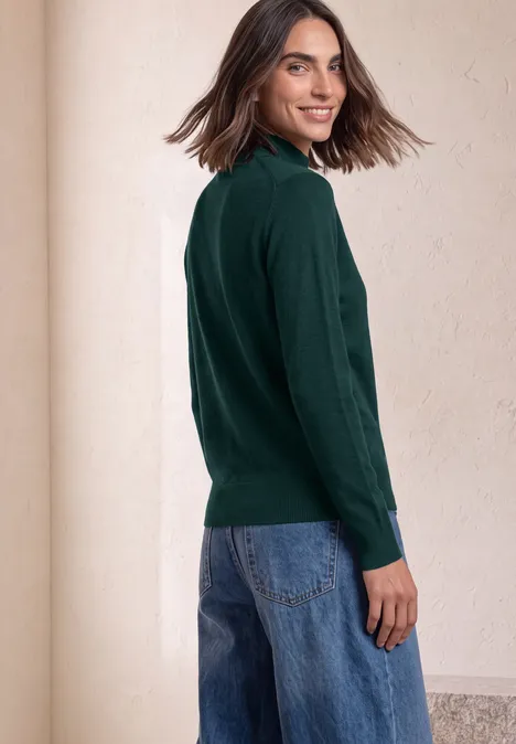 Basic sweter z golfem shadow teal Kobieta w ciemnozielonym swetrze i niebieskich jeansach spogląda przez ramię z uśmiechem.