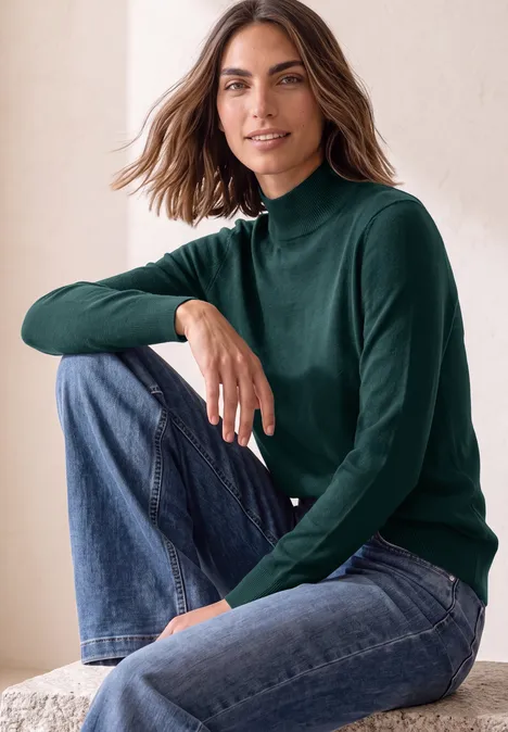 Basic sweter z golfem shadow teal Kobieta uśmiecha się, siedząc w dżinsach i zielonym golfie.