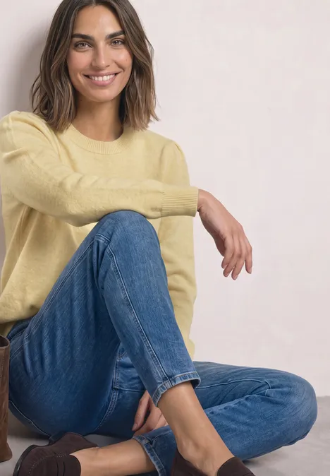 Pull à manches longues, col rond et fronces sunbeam yellow mel. Femme en pull jaune et jean bleu, assise détendue et souriante.
