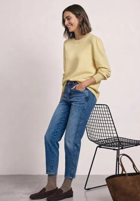 Pull à manches longues, col rond et fronces sunbeam yellow mel. Une femme porte un pull jaune, un jean bleu et des chaussettes marron, à côté d'une chaise en métal.
