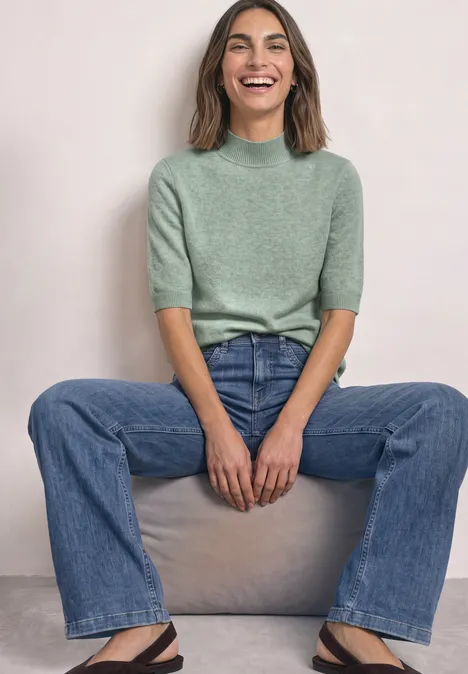 Kurzarm Pullover mit Turtle Neck eucalyptus green mel. Eine lächelnde Frau in graugrünem Pullover und Jeans sitzt entspannt auf einem Block vor neutralem Hintergrund.