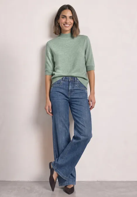 Kurzarm Pullover mit Turtle Neck eucalyptus green mel. Eine Frau mit grünem Pullover, weitem Jeans und schwarzen Schuhen lehnt lächelnd an einer hellen Wand.