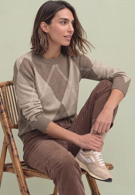 Eine Frau sitzt auf einem Rattanstuhl, trägt einen beigen Pullover mit Rautenmuster und braune Cordhose.