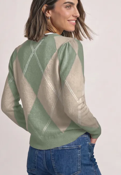 Vrouw in groen-beige argyle trui, glimlachend, kijkt over haar schouder.