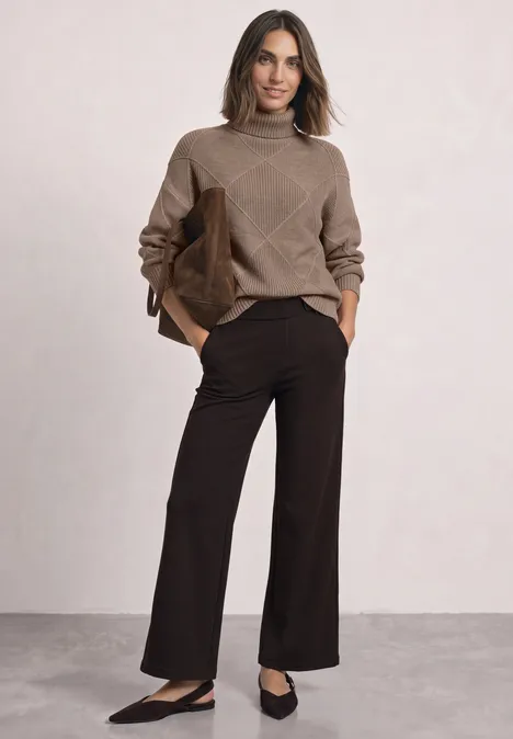 Frau trägt braunen Pullover und weite dunkelbraune Hosen, schwarze Ballerinas und hält braune Tasche.