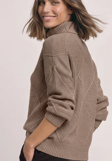 Eine lächelnde Frau trägt einen beigen, strukturierten Strickpullover mit Stehkragen und blickt über die Schulter.