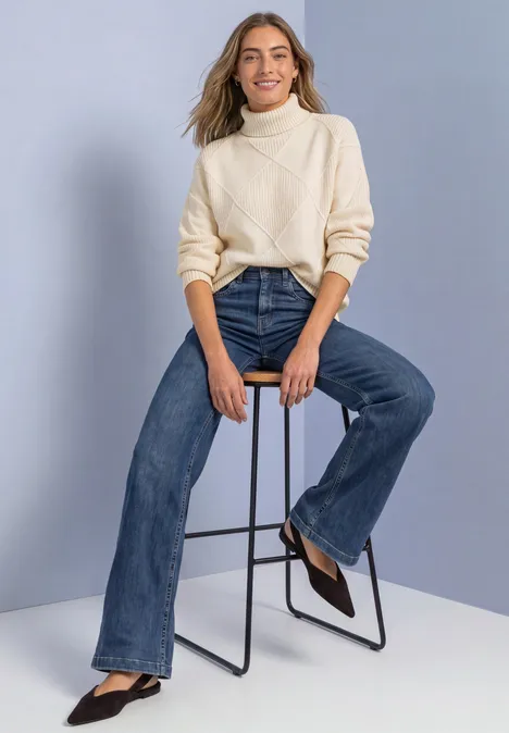 Frau in cremefarbenem Rollkragenpullover und weiten Jeans sitzt auf Hocker, trägt schwarze Pumps.