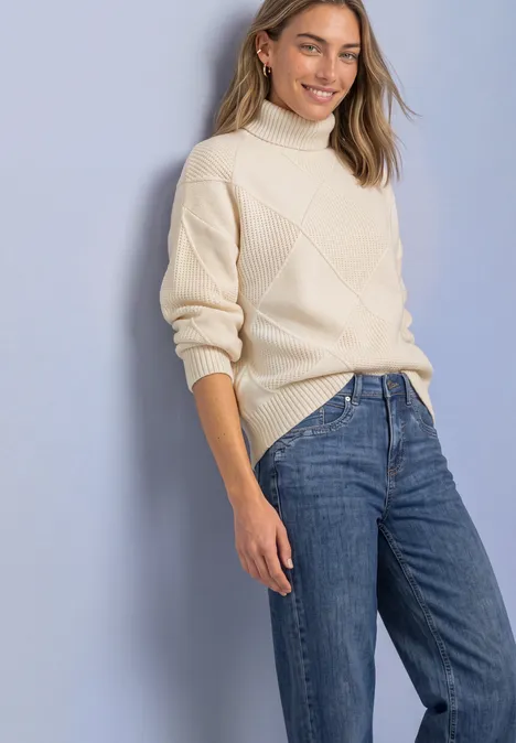 Frau in cremefarbenem Rollkragenpullover und blauen Jeans, lächelnd vor hellem Hintergrund.