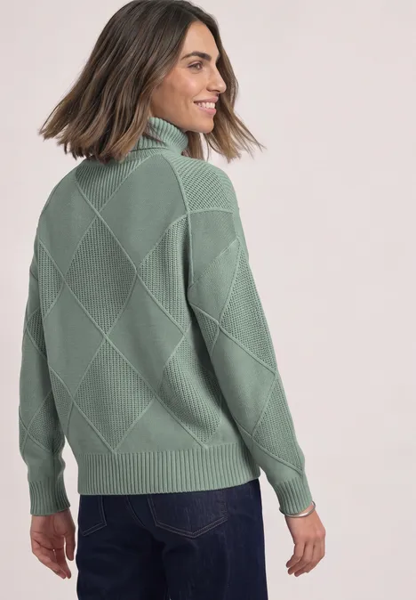 Frau sieht über die Schulter, trägt grünen Strickpullover mit Rautenmuster und Jeans.