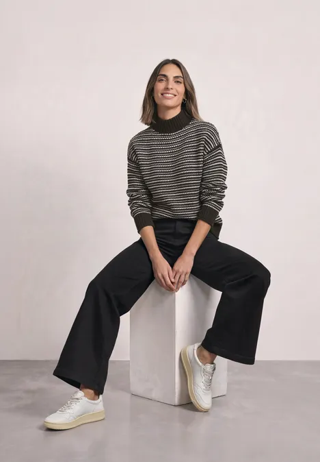Jonge vrouw in gestreepte trui, zwarte broek en witte sneakers zittend ontspannen op een witte blok.