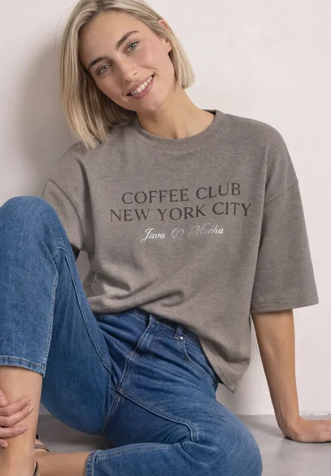 Frau sitzt lächelnd, trägt graues T-Shirt mit Aufschrift „Coffee Club New York City“ und blaue Jeans.