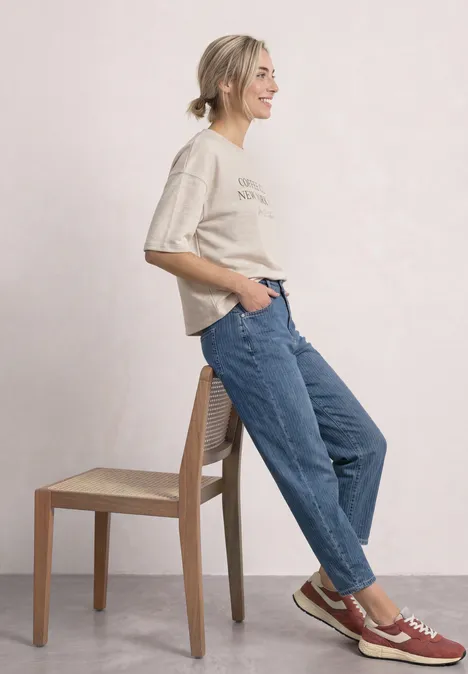 Eine Frau lehnt sich lässig an einen Stuhl, trägt T-Shirt, Jeans und Sneakers.