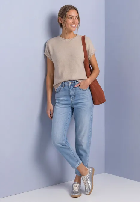 Pull sans manches à col rond light sepia sand Eine Frau lächelt vor violettem Hintergrund, trägt graues T-Shirt, Jeans, silberne Sneaker und eine braune Tasche.